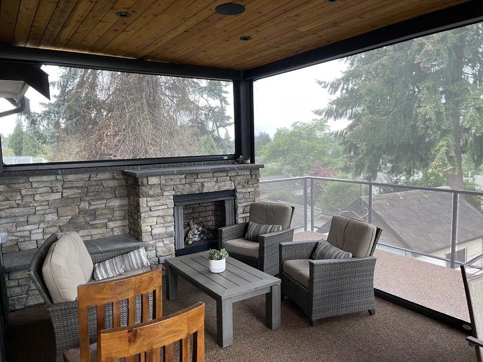 Black 5% openness motorized outdoor roller shades around a cosy covered patio with a stone fireplace and garden views