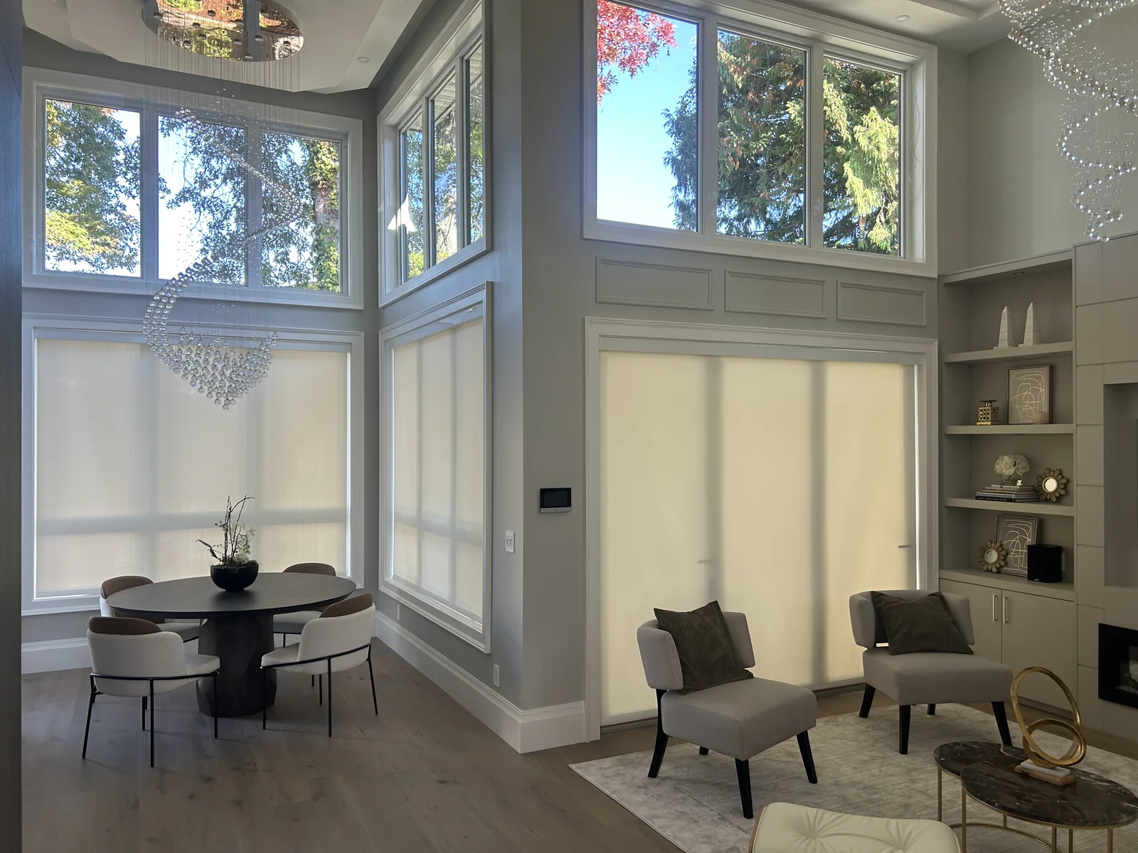 Light-filtering roller shades at the lower windows of a double-height living and dining room, with trees visible through the upper glazing