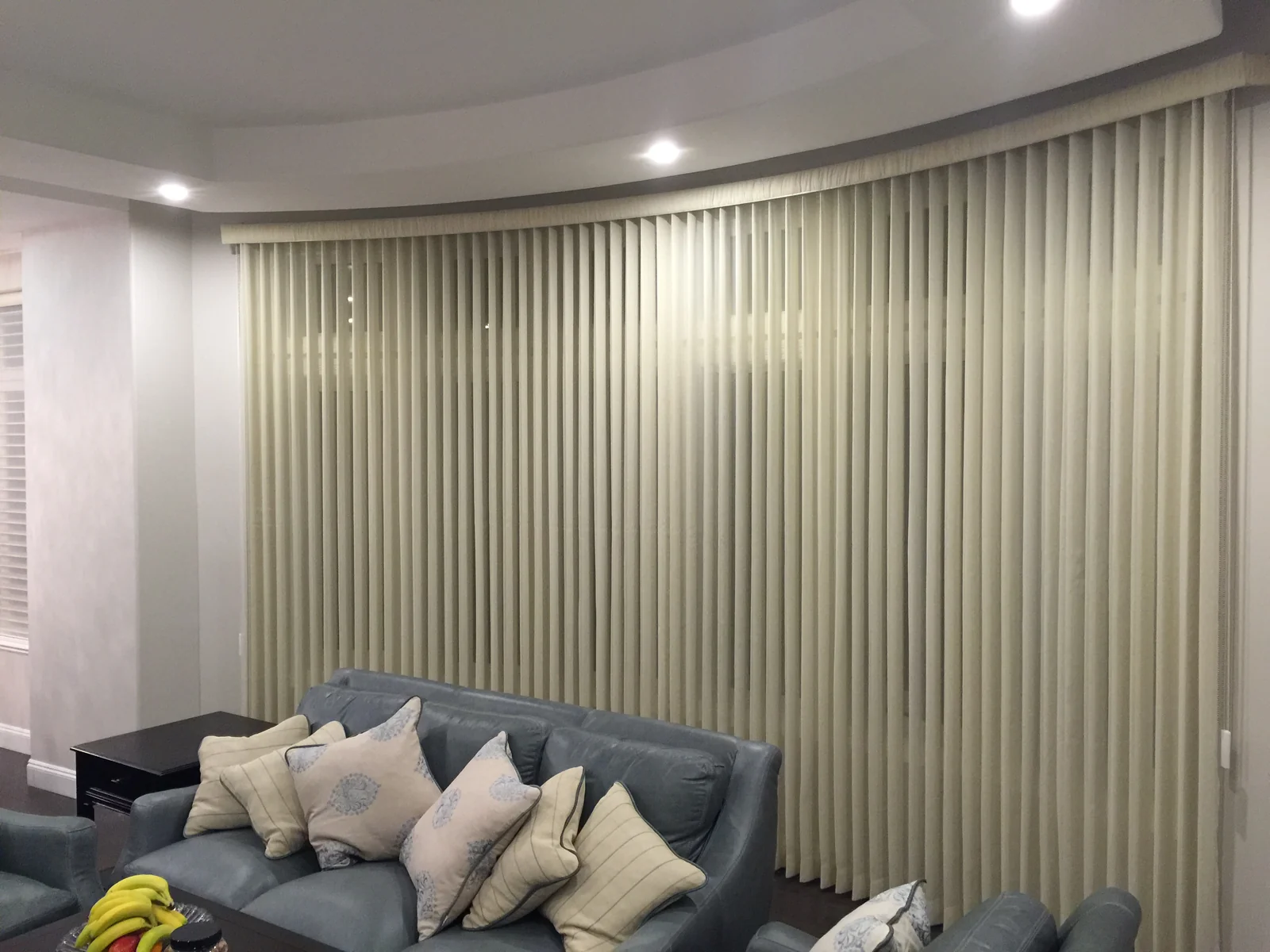 Sheer Vertical combines vertical blinds with a sheer fabric layer along a curved window wall behind a blue-grey sofa